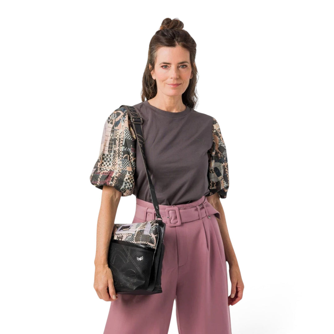 Woman wearing an Anekke crossbody bag with black and brown graphic patterns, styled with a grey top and pink trousers.
