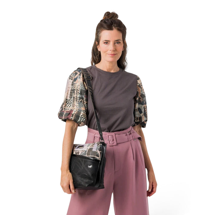Woman wearing an Anekke crossbody bag with black and brown graphic patterns, styled with a grey top and pink trousers.