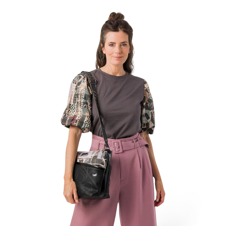 Woman wearing an Anekke crossbody bag with black and brown graphic patterns, styled with a grey top and pink trousers.